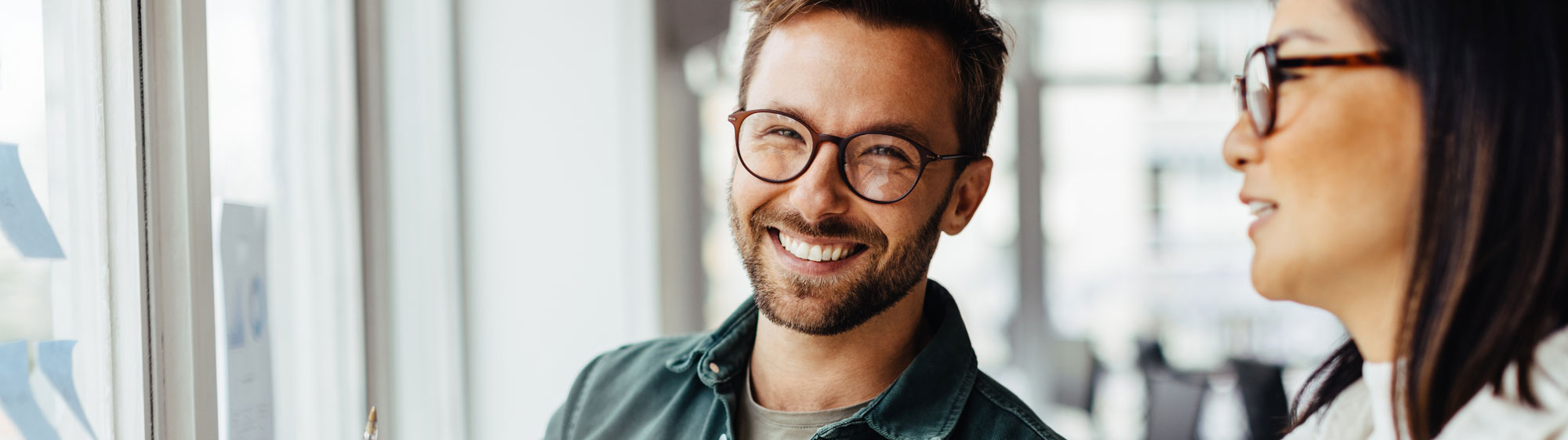 Ein Mann mit Brille im Büro blickt strahlend lächelnd in die Kamera während eine Frau neben ihm etwas äußert.